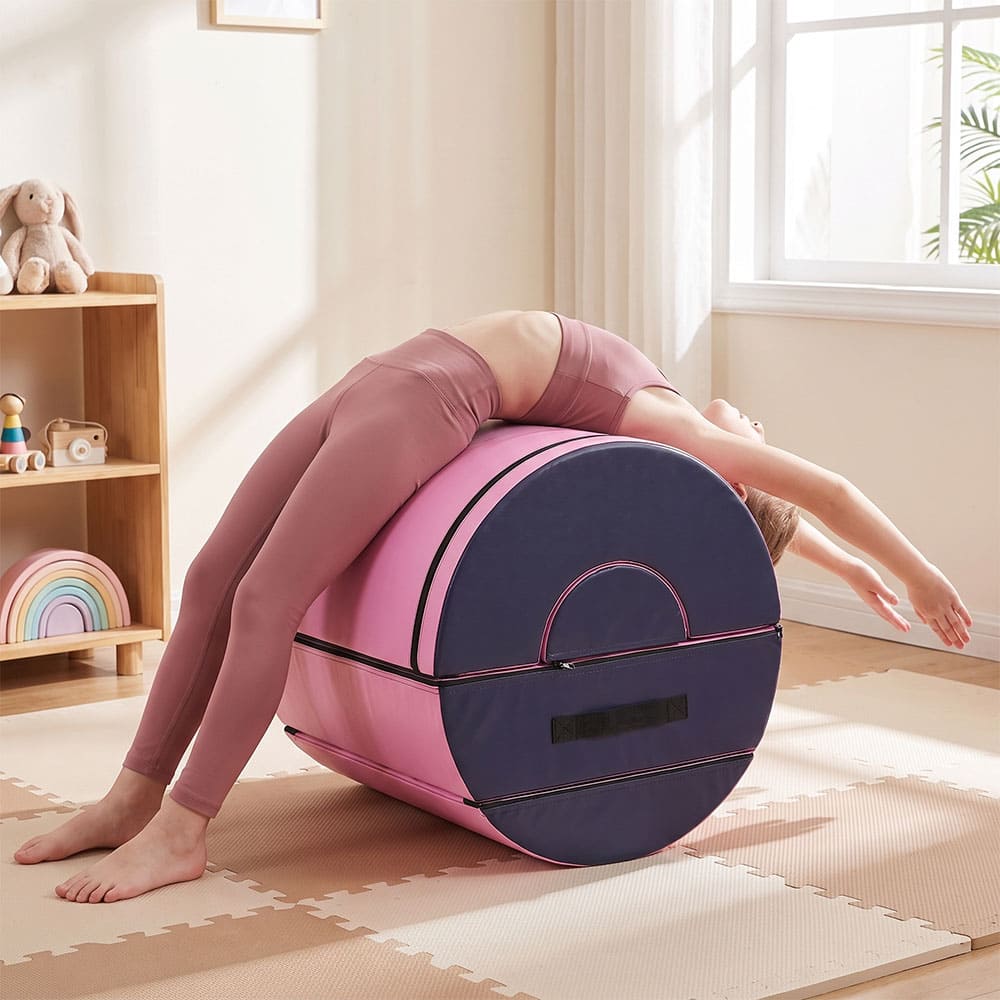 A child in a pink workout outfit performs a back stretch pose on a pink and purple roller in a bright, sunlit room with a wooden shelf and window.