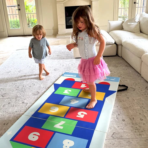 Two children playing on colorful numbered indoor hopscotch floor mat in living room
